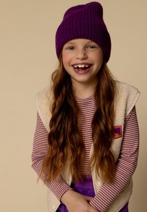 Kind dat een paarse geribbelde beanie, een gestreepte long-sleeve shirt en een fluffy crèmekleurige vest met een logo-patch draagt, en glimlacht met lang haar.
