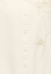 Tissu crème avec de petits boutons au centre et une broderie florale discrète en relief sur le côté droit.