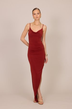 Femme en robe rouge sans manches, longue jusqu'au sol avec une fente latérale, sandales dorées, collier, boucles d'oreilles et bracelet, debout devant un fond beige.