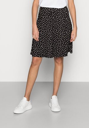 Mujer con falda negra hasta la rodilla con estampado abstracto blanco y marrón, zapatillas blancas y camiseta blanca, de pie sobre un suelo gris.