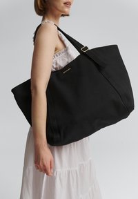 Zwarte canvas schoudertas met een grote, rechthoekige vorm en brede schouderbanden. Voorzien van goudkleurige "Hey Marly" branding en een gladde textuur.