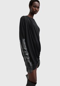 Abito nero drappeggiato con lunghi guanti di pelle lucida. L'abito presenta una vestibilità rilassata e una texture morbida, evidenziando una silhouette elegante.
