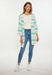 Proužkovaný cardigan v zelené, bílé a šedé barvě, s volným střihem, širokými rukávy a střední délkou, v kombinaci s modrými skinny džíny a bílými teniskami.