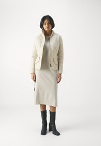 Jaqueta cream acolchoada com bolsos de botão, sobre um vestido bege até ao joelho. Botas pretas até ao joelho completam o conjunto. Design simples e limpo.