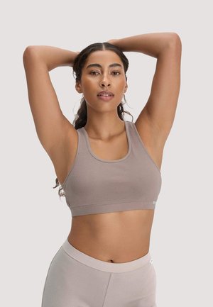 Vrouw met krullend haar in een taupekleurige geribbelde sport-bh en bijpassende broekjes, poserend met haar armen omhoog achter haar hoofd.