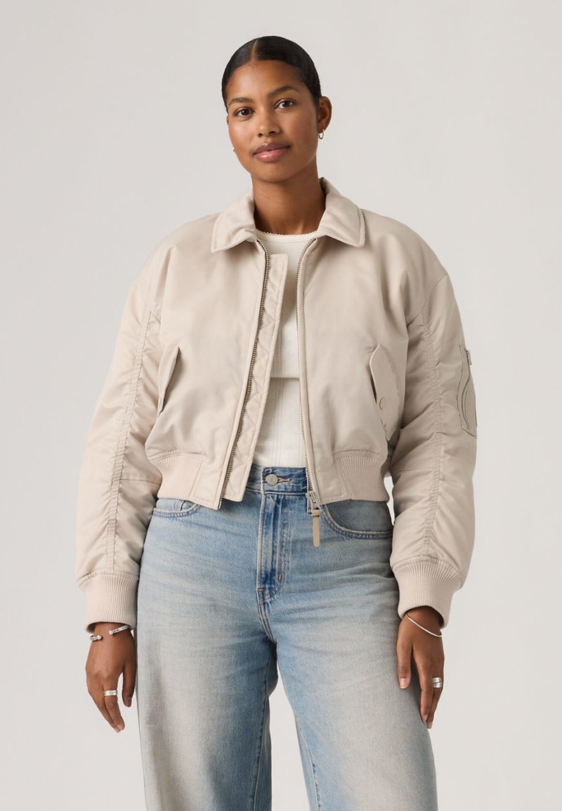Femme portant une veste bomber beige courte sur un haut blanc avec un jean taille haute bleu clair, debout devant un fond uni.