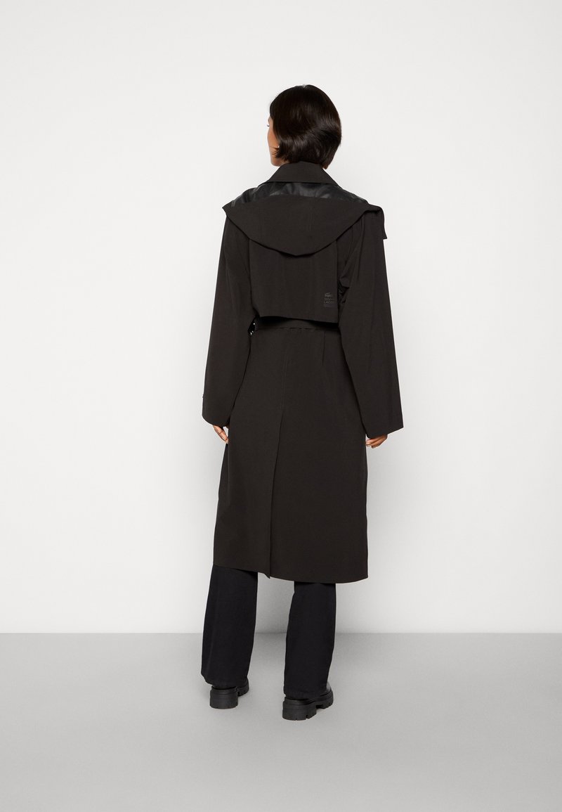 Cappotto nero in stile trench con design cropped, dotato di cappuccio e dettagli lucidi. Il tessuto è liscio con una silhouette strutturata.