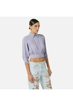 Donna con capelli pettinati all'indietro che indossa una camicetta lavanda corta con colletto e jeans chiari a vita alta con effetto consumato.