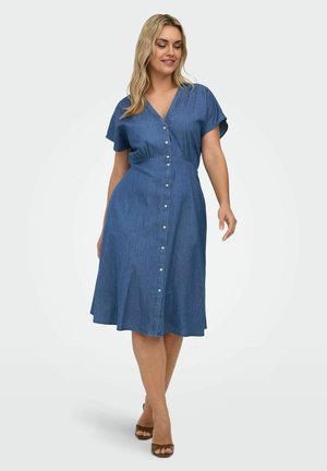 Kobieta idąca w niebieskiej sukience o długości do połowy łydki z guzikami z przodu, krótkimi rękawami i brązowymi sandałami na obcasie na białym tle.