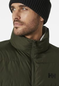 Groene pufferjack met een hoge kraag, ritsluiting en zwart logo op de borst. Gekleed met een zwarte geribbelde beanie.