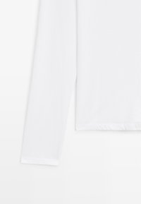 Wit shirt met lange mouwen en een ronde hals, deels zichtbaar van de mouw tot aan de zoom, op een witte achtergrond.