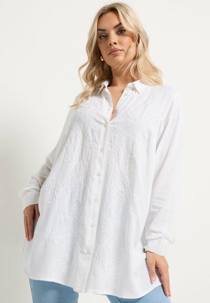 Blonde woman wearing loose white button-up shirt with floral embroidery, blue jeans, standing against plain light background.