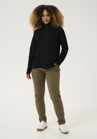 Schwarzer gerippter Pullover mit hohem Kragen und halbem Reißverschluss, kombiniert mit olivgrünen Slim-Fit-Hosen und weißen Sneakers.