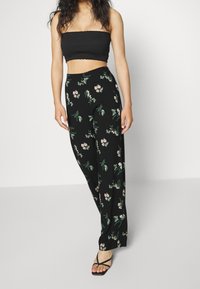 Žena nosící černé květinové široké kalhoty s černým bezramenným crop topem a černými sandály na podpatku, stojící proti bílé zdi.