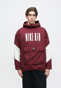 Casaco bordeaux com capuz, apresentando um grande logótipo "NIKE AIR" em branco, mangas brancas em contraste, e um bolso frontal com detalhe de etiqueta.