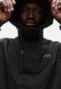 Μαύρο αδιάβροχο μπουφάν με ψηλό γιακά, ρυθμιζόμενα κορδόνια και λογότυπο στο στήθος. Λεπτή υφή και εφαρμοστή σχεδίαση.