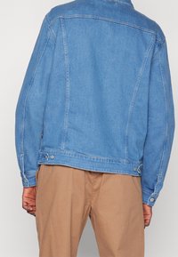 Person wearing a blue denim jacket and light brown pants, shown from the back against a plain light background.