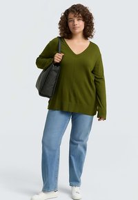 Maglione verde lavorato a maglia con scollo a V e maniche lunghe, abbinato a jeans azzurri e sneakers bianche. La modella porta una borsa nera.