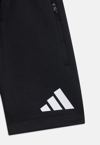 Čierne atletické šortky s hladkou textúrou, vybavené zipsovým vreckom a bielym logom s tromi pruhmi na leme.