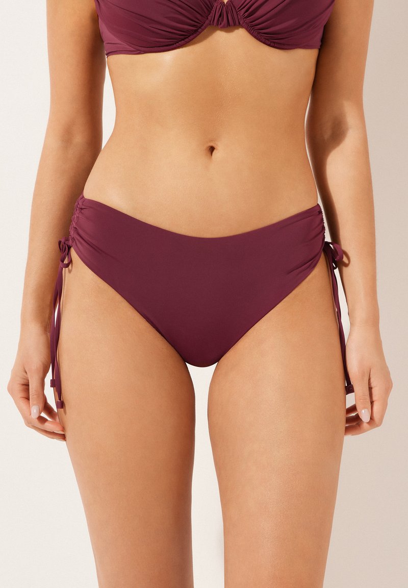 Žena oblečená v tmavě fialových bikini kalhotkách s nastavitelnými bočními šňůrkami, zobrazená od pasu po horní část stehen na jednobarevném pozadí.