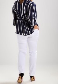 Donna di spalle che indossa una maglietta a righe blu navy e bianche con una cintura annodata, pantaloni bianchi e scarpe col tacco nere con cinturini alla caviglia.