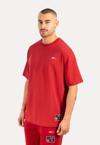 Camiseta de algodón roja con cuello redondo, mangas cortas y un pequeño logo blanco. Presenta un corte relajado y elementos de diseño minimalistas.