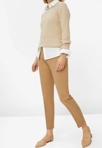 Beige strikket sweater lagdelt over en hvid skjorte, parret med brune tapered bukser og nude slip-on flade sko. Enkel design og neutrale farver.
