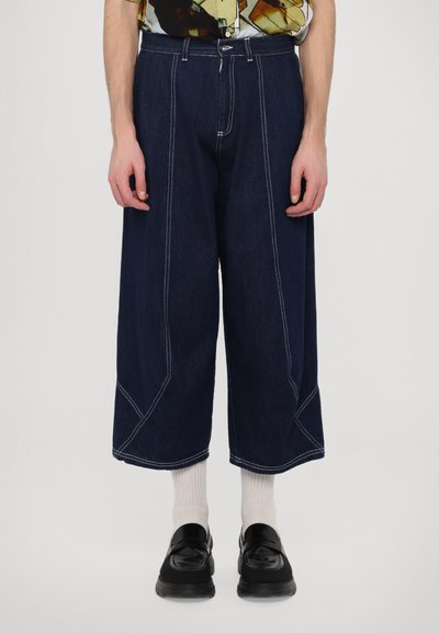 Mørkeblå denim beskåret bukser med brede ben, der har kontrasterende hvide syninger og en frontknaplukning. Båret med sorte sko og hvide sokker.