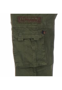 Poche d’un pantalon cargo vert olive avec rabat et écusson "ALPHA" cousu ainsi qu’une petite étiquette en tissu noire avec logo orange sur le côté.