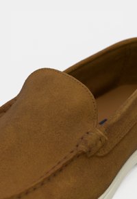 Zapato de ante marrón tipo slip-on con una textura suave, punta redondeada, sutura sutil y una suela de goma blanca. Logotipo grabado en el lateral.