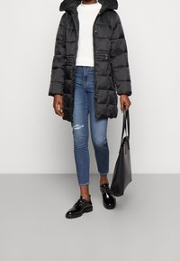 Abrigo de plumas negro con capucha, cierre de cremallera y detalle ajustado en la cintura. Lleva una blusa blanca debajo y lo combina con unos jeans azules desgastados y zapatos negros.