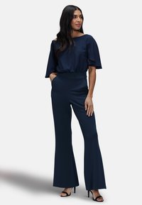 Navyblauwe wijd uitlopende broek en top met korte mouwen van een gladde stof. Kenmerkt zich door een brede tailleband en open mouwen. Gemaakt met hoge sandalen.