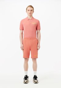 Conjunto de camiseta polo y pantalones cortos en color coral; material de algodón, corte relajado, detalle de logotipo en la camiseta y en los pantalones cortos, combinado con zapatillas de deporte con un diseño estampado.