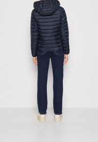 Persona che indossa una giacca a cappuccio trapuntata blu navy, jeans dritti blu scuro e stivali spessi color avorio, in piedi davanti a uno sfondo semplice.