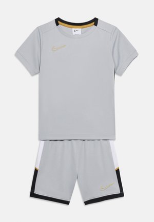 Svetlosivé športové oblečenie Nike s krátkymi rukávmi a šortkami, vybavené zlatými logami Nike Swoosh a čiernymi, bielymi a zlatými akcentmi.