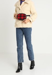Veste en fausse fourrure crème, jean denim bleu, bottes chelsea en cuir verni noir et sac banane à carreaux rouges. Design simple et tissu texturé.