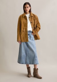 Braune Wildlederjacke mit großen Taschen, weißes Hemd und hellblaue Jeans-Maxiröhre. Kombiniert mit taupefarbenen spitz zulaufenden Ankle Boots.