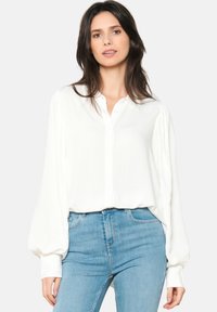 Camicia bianca a maniche lunghe con chiusura a bottoni, texture morbida e maniche arricciate, abbinata a jeans aderenti azzurri.
