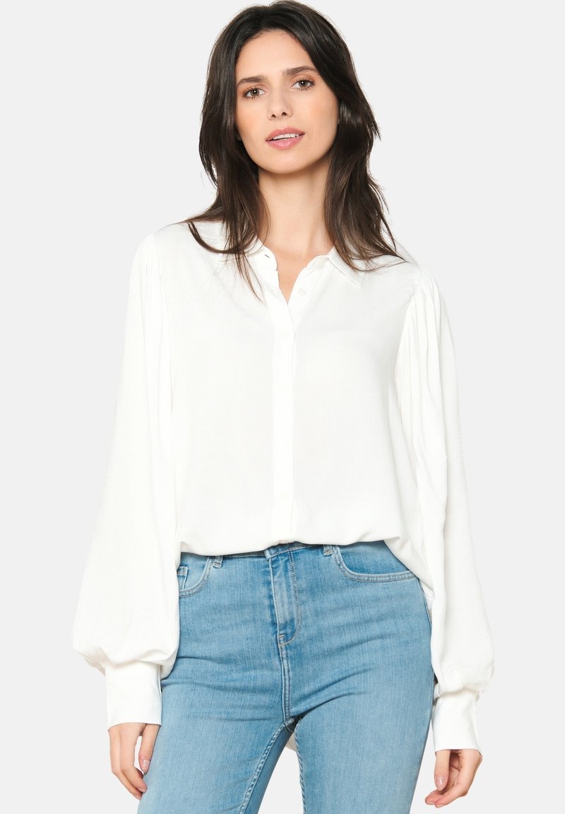 Camicia bianca a maniche lunghe con chiusura a bottoni, texture morbida e maniche arricciate, abbinata a jeans aderenti azzurri.