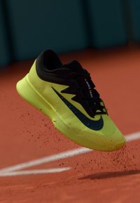 Παπούτσι τένις Nike με φωσφορίζον κίτρινο επάνω μέρος, μαύρες λεπτομέρειες και υφασμένο πέλμα, κρεμασμένο πάνω από μια κόκκινη επιφάνεια γηπέδου, διασπείροντας σκόνη.