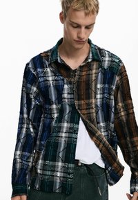 Plaid shirt in green, blue, and brown tones, featuring a button-down design, textured fabric, and frayed edges. Worn over a white t-shirt.