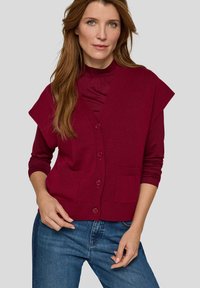 Bordeauxroter Cardigan mit kurzen Ärmeln, Knopfverschluss und Fronttaschen, kombiniert mit einem eng anliegenden Top, getragen zu blauen Jeans. Glatte Textur.