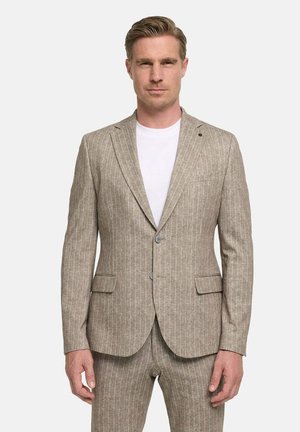 Man wearing beige pinstripe suit jacket and matching pants over a white t-shirt, standing against a plain light background.