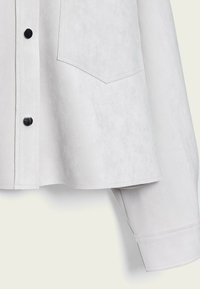 Chemise en daim gris clair, avec une patte de boutonnage, des boutons en métal noir et une poche poitrine, dotée d'un ourlet droit et de manches à revers.