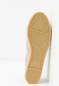 Semelle d'une espadrille grise avec une semelle extérieure en caoutchouc brun clair, taille 37, marquée "Fabriquée en Espagne", positionnée debout contre une surface blanche.