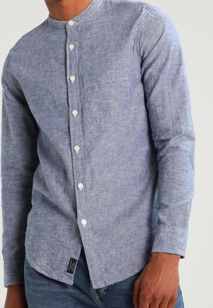 Man wearing a light blue long-sleeve button-up shirt with a band collar and chest pocket, paired with blue jeans.