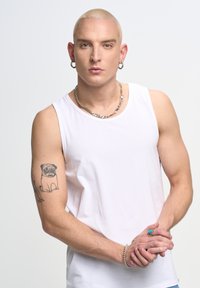 Balta bez piedurknēm krekls, sudraba ķēdes kaklarota, pug tetovējums uz kreisās rokas, vīrieša figūra ar īsiem blondiem matiem, rokas saliktas kopā.