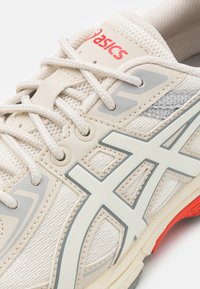 Chaussure de course beige avec tige en mesh, accents gris et superpositions synthétiques blanches. Présente un logo rouge et une semelle extérieure texturée pour l'adhérence.