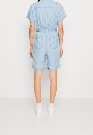 Person iført lyseblå denim kortærmet heldragt med to baglommer, opkørte shorts, hvide sokker og hvide sneakers, set bagfra.
