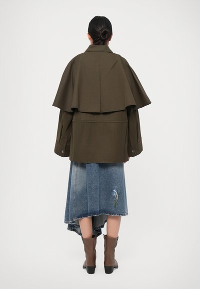 Olive green layered jacket with a cape-like design, paired with a blue denim skirt featuring an iris embroidery and brown ankle boots.